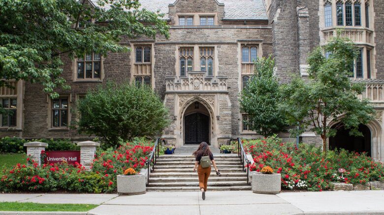 A person with tan pants and a black short and backpack walks toward a McMaster University building. They have long hair. Trees and flowers in bloom are shown around the building, as well as a sign that reads "University Hall."