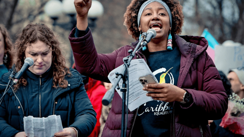 Image description: an Afro- and Indo-Caribbean trans woman with trans flag earrings in a dark red jacket and black sweater speaking into a microphone.